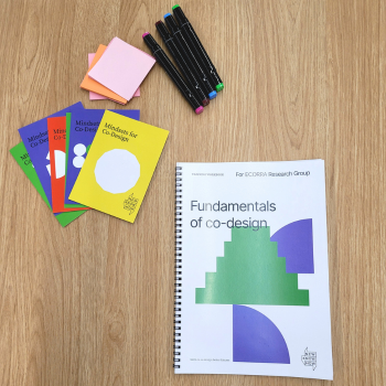 "Fundamentals of Co-design" spiral-bound workbook beside colourful "Mindsets for Co-Design" cards and markers on a wooden table.